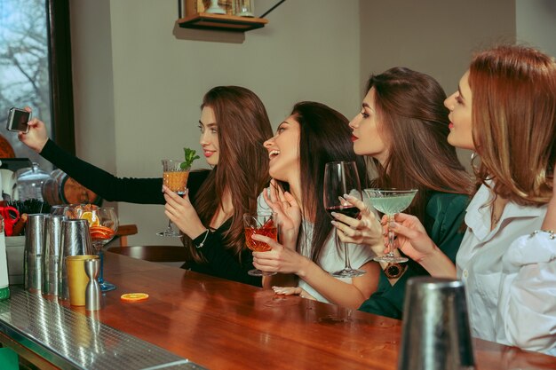 Amigas tomando una copa en el bar. Están sentados en una mesa de madera con cócteles. Llevan ropa informal.