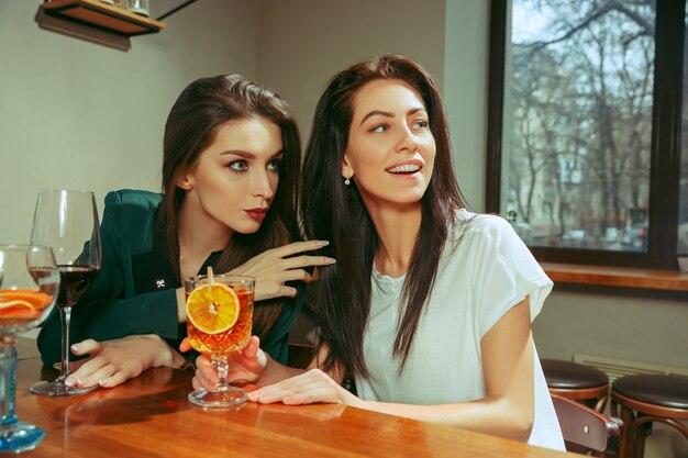 Amigas tomando una copa en el bar. Están sentados en una mesa de madera con cócteles. Llevan ropa informal.