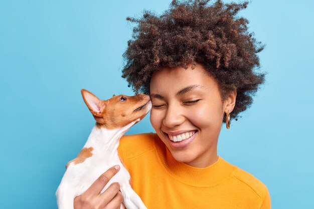 Amigo de la familia. Primer plano de feliz mujer de pelo rizado juega con perro expresa emociones positivas como animales. Pequeño cachorro de pedigrí lame la cara del propietario. Mascota adoptada. Tiernos sentimientos sinceros