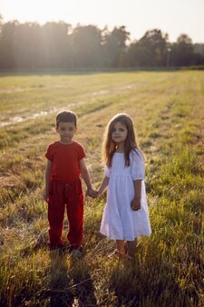 Amigo, un niño con ropa roja y una niña con un vestido blanco caminan por un campo segado al atardecer en verano