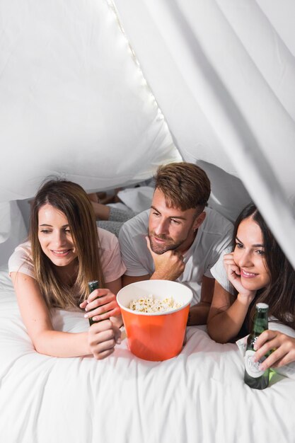 Amigos acostados en la cama con palomitas de maíz y bebidas viendo la televisión