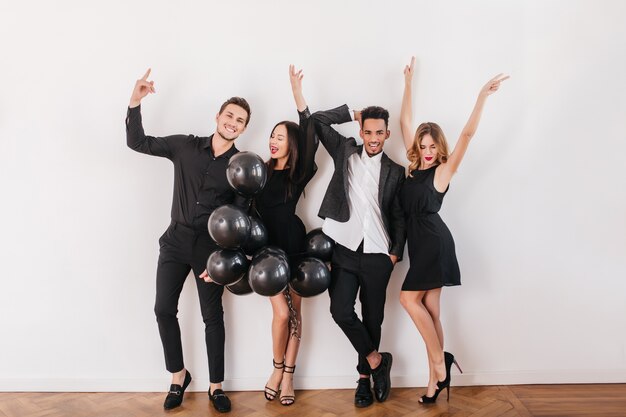 Amigos alegres bailando en la pared blanca con globos negros durante la fiesta en casa