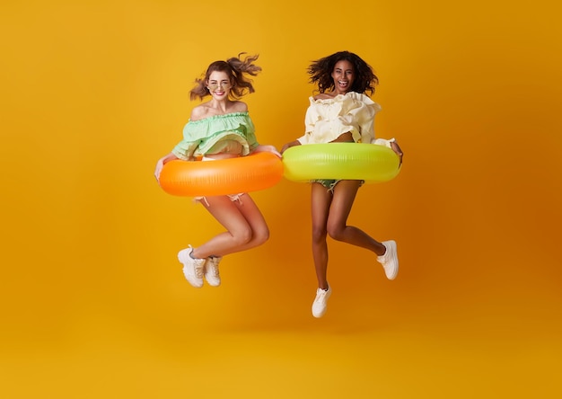 Amigos alegres mujer vestida con ropa de verano saltando con anillo de goma sobre fondo amarillo