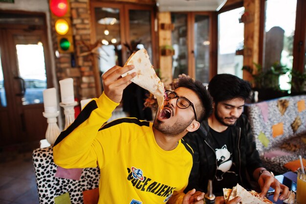 Amigos asiáticos chicos comiendo pizza durante la fiesta en la pizzería Gente india feliz divirtiéndose juntos comiendo comida italiana y sentados en el sofá