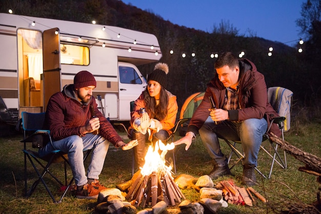 Amigos cercanos bebiendo cerveza juntos en las montañas y calentando sus manos alrededor de la fogata. Autocaravana retro con bombillas.