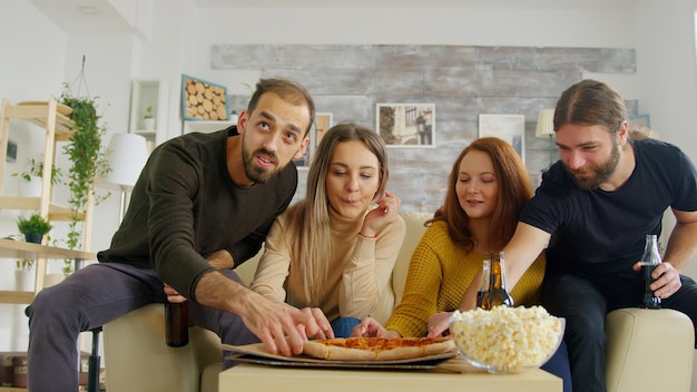 Los amigos cercanos ven la televisión en la sala de estar animándose mientras ven un partido de fútbol en el sofá. Amigos comiendo bocadillos.