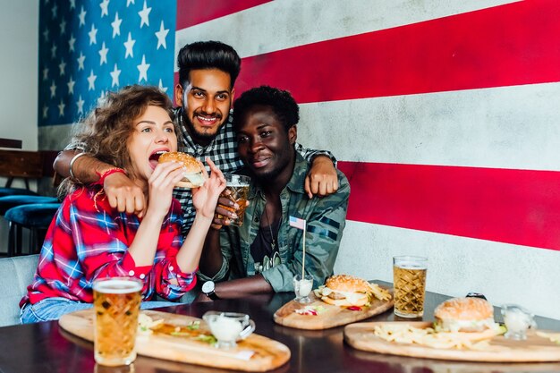 Amigos felices abrazándose, comiendo hamburguesas, hablando y sonriendo mientras pasan tiempo juntos en la cafetería.