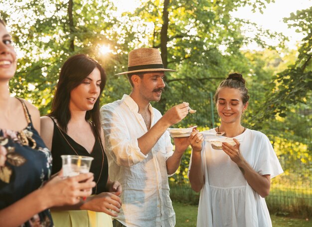 Amigos felices comiendo y bebiendo cervezas en la cena de barbacoa al atardecer