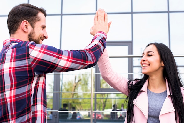 Amigos felices jovenes del hombre y de la mujer que dan el edificio de cristal cercano alto cinco