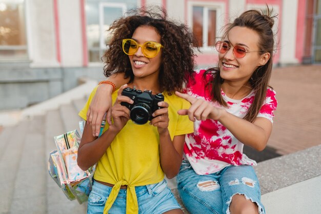 Amigos felices de las niñas sonrientes sentados en la calle con cámara de fotos, mujeres divirtiéndose juntos