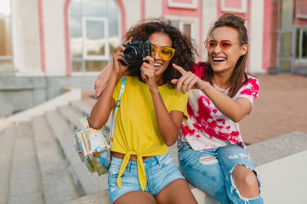 Amigos felices de las niñas sonrientes sentados en la calle con cámara de fotos, mujeres divirtiéndose juntos