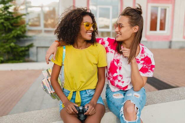 Amigos felices de las niñas sonrientes sentados en la calle con cámara de fotos, mujeres divirtiéndose juntos