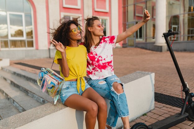 Amigos felices de las niñas sonrientes sentados en la calle tomando fotos selfie en el teléfono móvil, mujeres divirtiéndose juntos