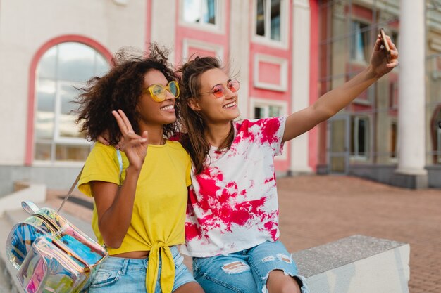 Amigos felices de las niñas sonrientes sentados en la calle tomando fotos selfie en el teléfono móvil, mujeres divirtiéndose juntos