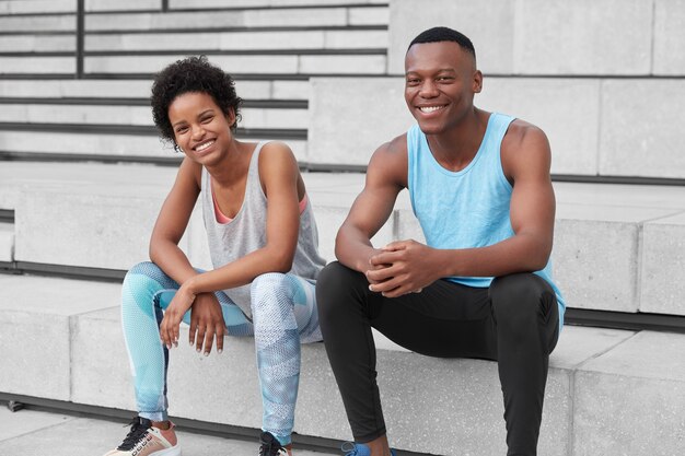 Amigos felices de piel oscura hacen ejercicio cardiovascular en un entorno urbano, descansan en las escaleras, usan chalecos sueltos, leggings, zapatillas de deporte, tienen expresiones alegres, logran los resultados necesarios, quieren estar sanos y en forma