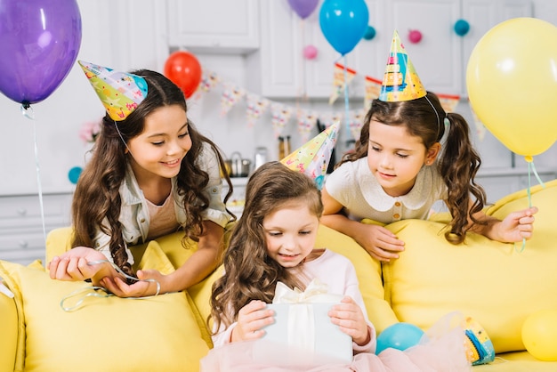 Amigos felices que miran a la muchacha del cumpleaños que abre el presente en casa