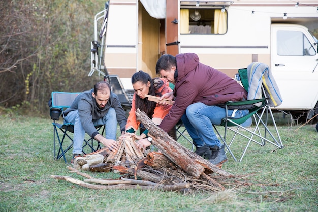 Amigos hipster haciendo juntos una fogata en el bosque de montaña. Amigos acampando con autocaravana retro.