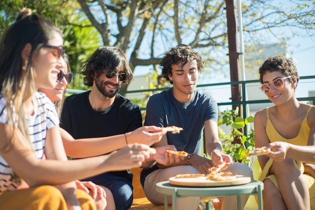 Amigos hombres y mujeres pacíficos sentados a la mesa y comiendo pizza en la fiesta. Jóvenes de diferentes nacionalidades con ropa informal sentados en el techo de la terraza, comiendo pizza. Fiesta, concepto de amistad