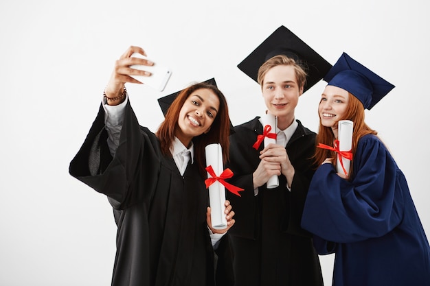 Amigos lindos graduados sonrientes sosteniendo diplomas haciendo selfie.