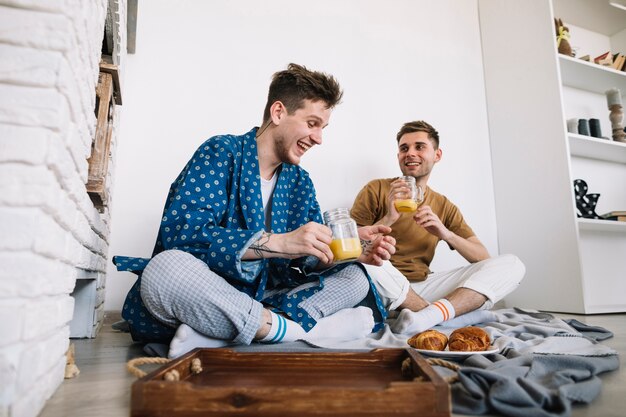 Amigos masculinos alegres que disfrutan del desayuno sabroso que se sienta en piso en casa