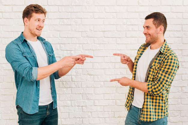 Amigos masculinos felices que se oponen a la pared blanca que se señalan los dedos el uno al otro