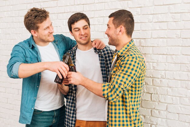 Amigos masculinos felices que se oponen a la pared blanca que tuesta las botellas de cerveza