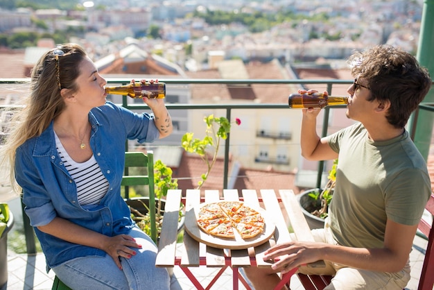 Amigos masculinos y femeninos relajados sentados a la mesa en la fiesta. Jóvenes con ropa informal sentados en el techo de la terraza, hablando, comiendo pizza y bebiendo cerveza. Comunicación, concepto de amistad