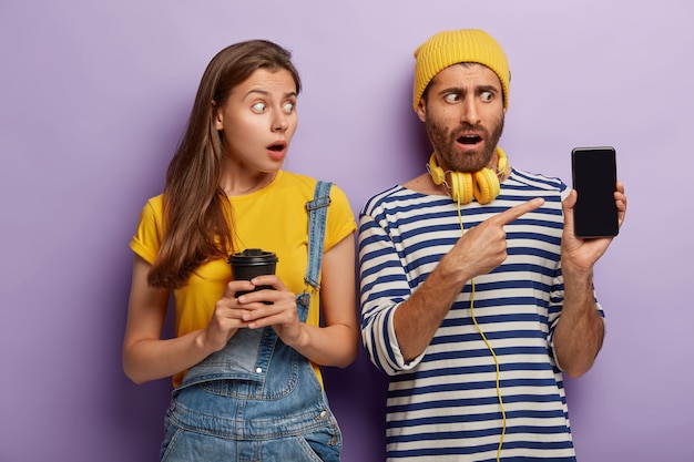 Amigos masculinos y femeninos sorprendidos señalan la pantalla del teléfono inteligente, muestran la pantalla de la maqueta, la mujer sostiene el café para llevar, vestida con un mono de mezclilla, se paran uno al lado del otro en el estudio