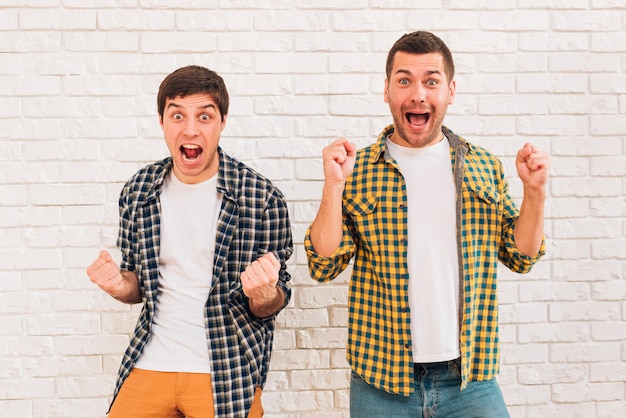 Amigos masculinos jovenes emocionados que se oponen a la pared de ladrillo blanca que aprieta su puño