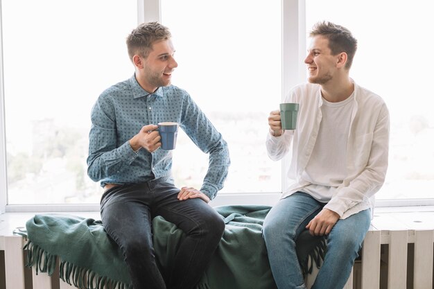 Amigos masculinos sonrientes que se sientan cerca de la ventana que come café