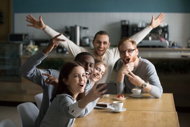 Amigos milenarios divertidos que toman grupo selfie en teléfono inteligente en café