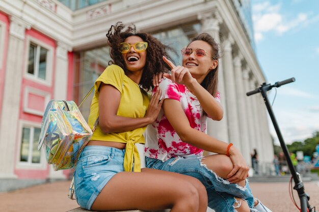 Amigos de las muchachas jóvenes felices con estilo colorido que sonríen sentados en la calle, mujeres divirtiéndose juntas