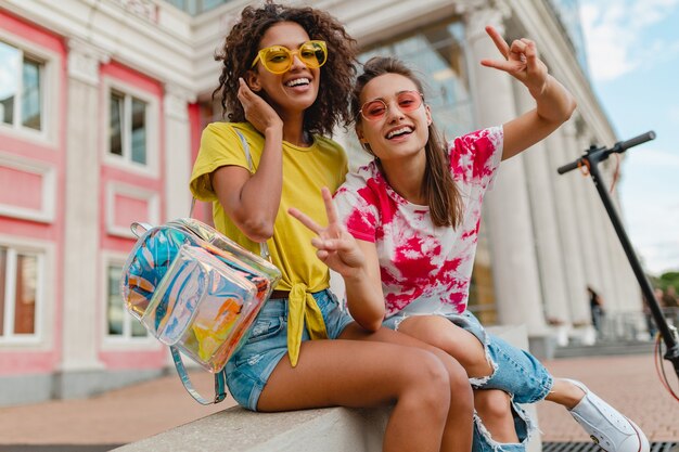 Amigos de las muchachas jóvenes felices con estilo colorido que sonríen sentados en la calle, mujeres divirtiéndose juntas