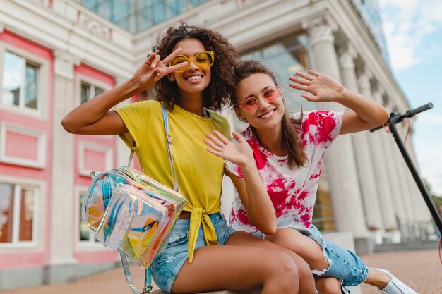 Amigos de las muchachas jóvenes felices con estilo colorido que sonríen sentados en la calle, mujeres divirtiéndose juntas