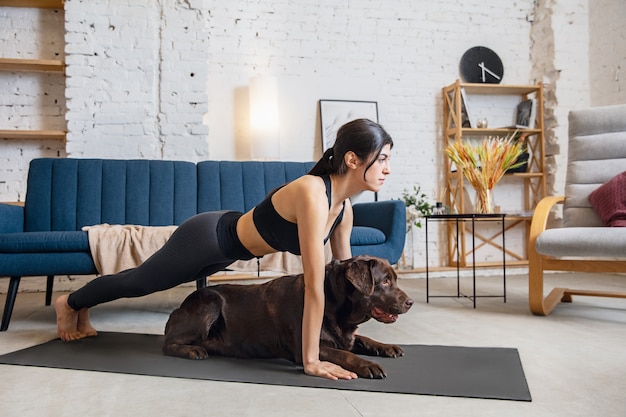 Amigos. Mujer joven trabajando en casa, haciendo ejercicios de yoga con el perro. Hermosa mujer estirando, practicando. Bienestar, bienestar, salud, salud mental, concepto de estilo de vida.