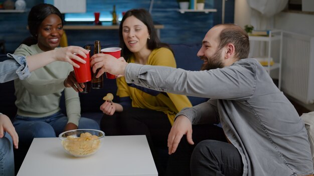 Amigos multiculturales socializando mientras están sentados en el sofá en la sala de estar a altas horas de la noche bebiendo cerveza e ...