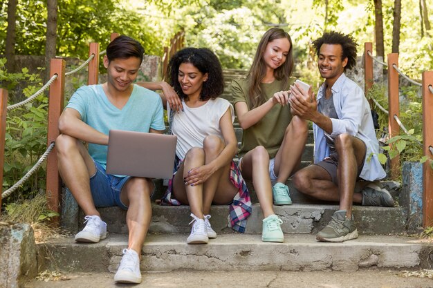 Amigos multiétnicos estudiantes al aire libre usando teléfono móvil y computadora portátil