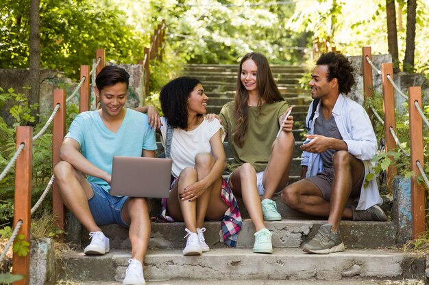 Amigos multiétnicos estudiantes al aire libre usando teléfono móvil y computadora portátil