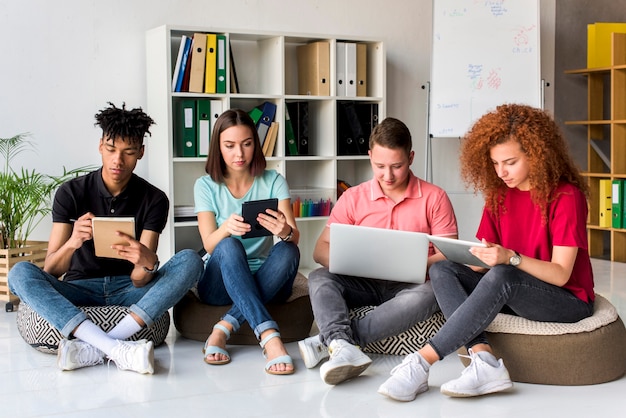 Amigos multirraciales que estudian con aparatos electrónicos y libros sentados en la sala de estudio