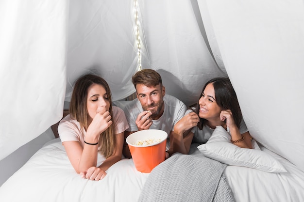 Amigos que mienten en la cama acogedora blanca que come palomitas de maíz