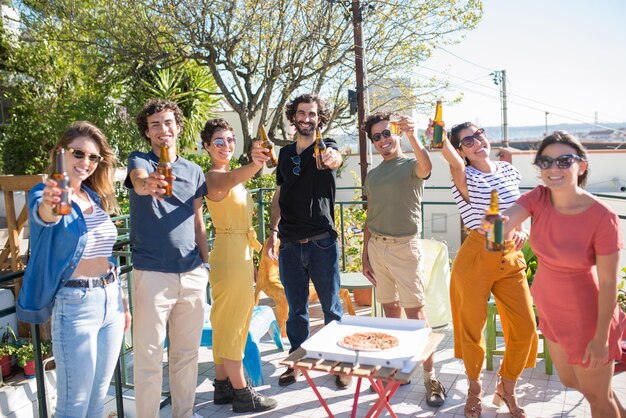 Amigos radiantes chocando botellas de cerveza. Gente de diferentes nacionalidades levantando botellas a la cámara, riéndose en un día soleado. Fiesta, concepto de amistad