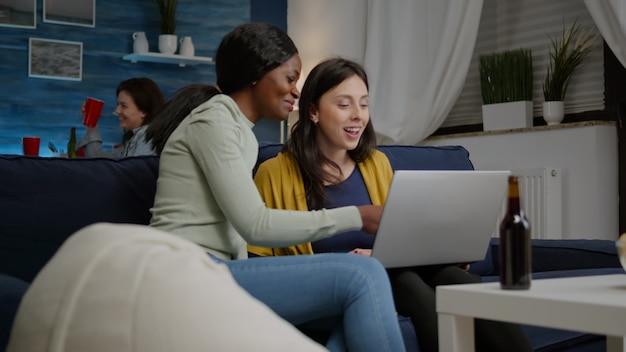 Amigos de raza mixta hablando mientras miran series de videos divertidos en la computadora portátil sentado en el sofá en la sala de estar ...