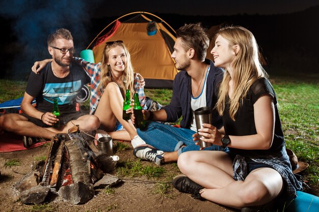 Amigos sentados cerca de la hoguera, sonriendo, hablando, descansando, bebiendo oso