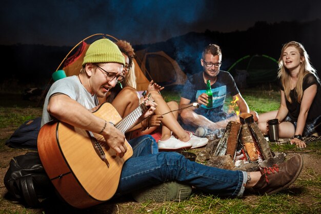 Amigos sentados cerca de la hoguera, sonriendo, hablando, descansando, tocando la guitarra