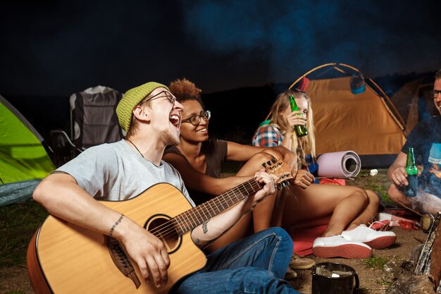 Amigos sentados cerca de la hoguera, sonriendo, hablando, descansando, tocando la guitarra