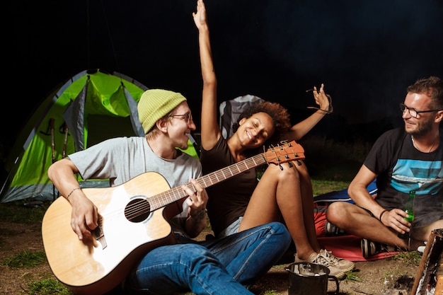 Amigos sentados cerca de la hoguera, sonriendo, hablando, descansando, tocando la guitarra