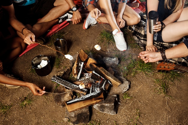 Amigos sentados cerca de la hoguera, sonriendo, tocando la guitarra Camping grill malvavisco.