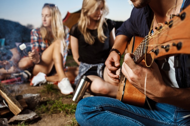 Amigos sentados cerca de la hoguera, sonriendo, tocando la guitarra Camping grill malvavisco.