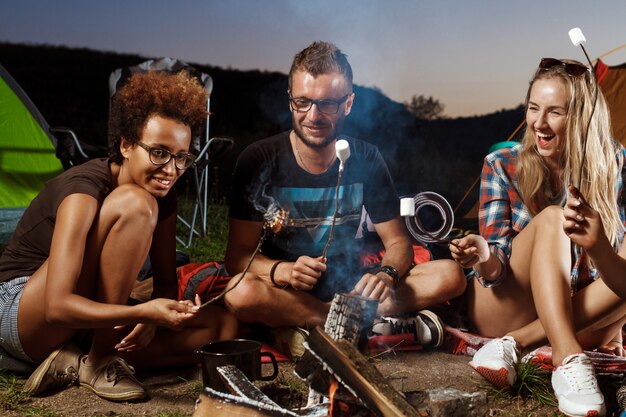 Amigos sentados cerca de la hoguera, sonriendo, tocando la guitarra Camping grill malvavisco.