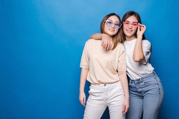 Amigos para siempre. Dos amigas encantadoras lindas en gafas de sol posando con una sonrisa aislada en la pared azul